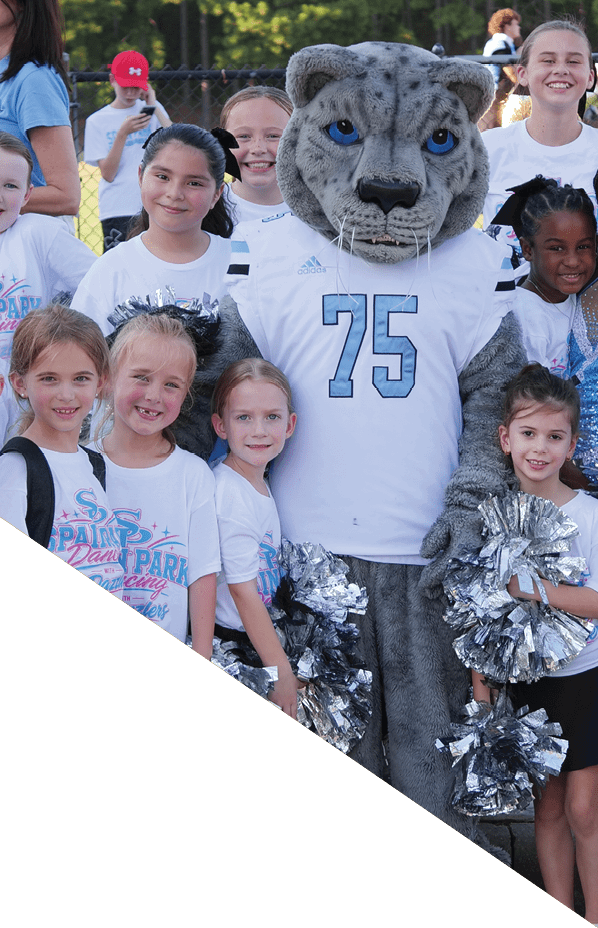 A group of children wearing matching white shirts pose happily around a person in a large gray animal mascot costume with blue eyes and a white jersey numbered 75. Description generated by AI