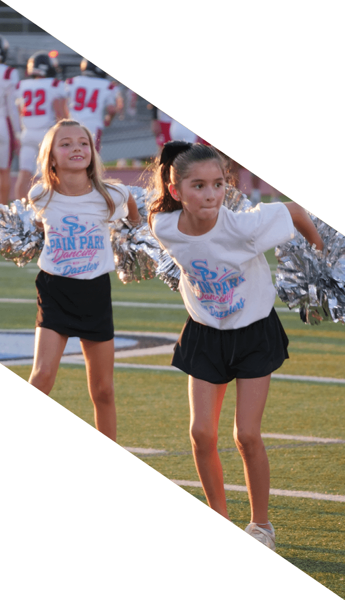 Children in matching cheerleading outfits perform with silver pom-poms on a sports field while a football team stands in the background. Description generated by AI