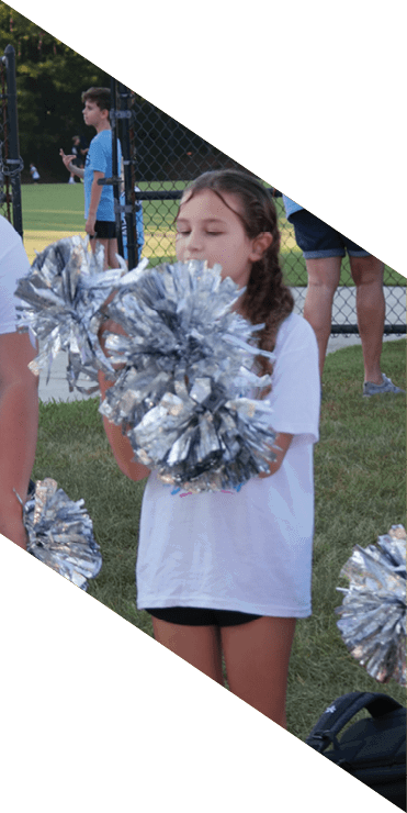 Two people wearing white shirts with “Spain Park Dance Team" printed on the back are holding silver pom-poms near a fenced sports field. Description generated by AI