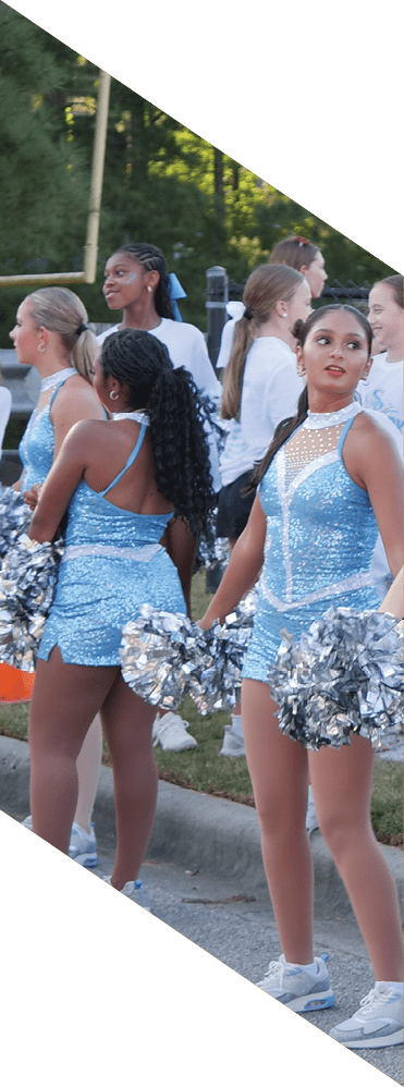 People in sparkly blue cheerleading outfits hold silver pom-poms while standing outdoors near a grassy area. Description generated by AI