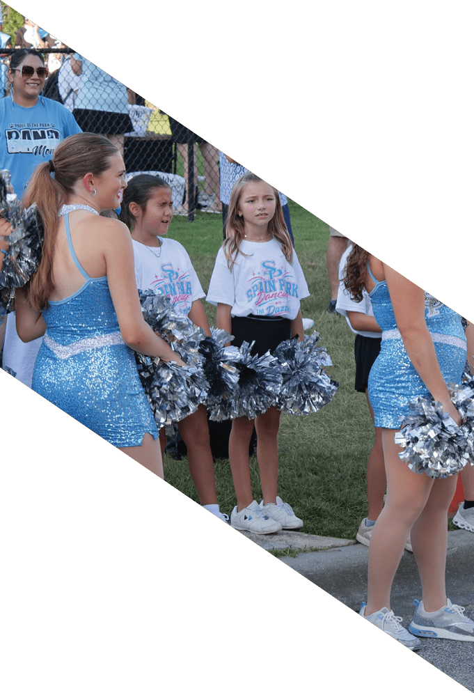 People in blue sparkly cheerleading outfits and white t-shirts holding silver pom-poms stand together on grass near a sidewalk. Description generated by AI