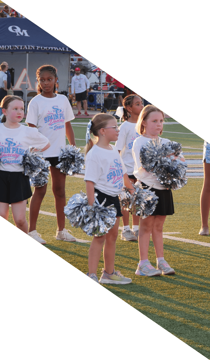People wearing matching white shirts and black skirts are standing on a sports field holding silver pom-poms, likely preparing for a cheerleading routine. Description generated by AI
