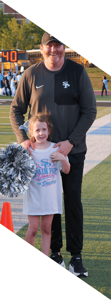 A child wearing a white shirt with colorful text holds silver pom-poms while standing in front of an adult on a sports field. Description generated by AI