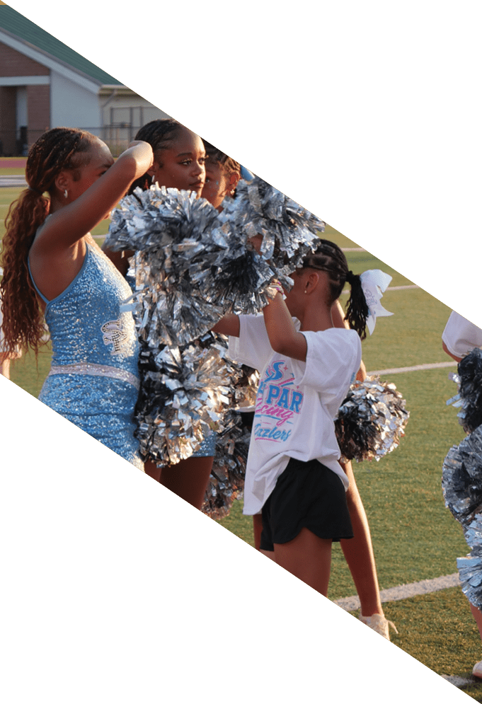 Children in cheerleading outfits and t-shirts are holding pom-poms and standing on a sports field during an outdoor event. Description generated by AI