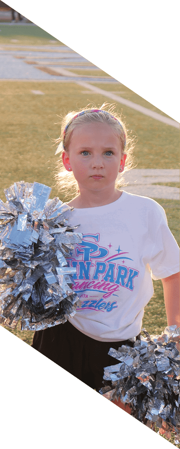 Two children wearing matching white t-shirts with “Spin Park Dancing The Dazzlers" printed on them hold silver pom-poms while standing on a sports field. Description generated by AI