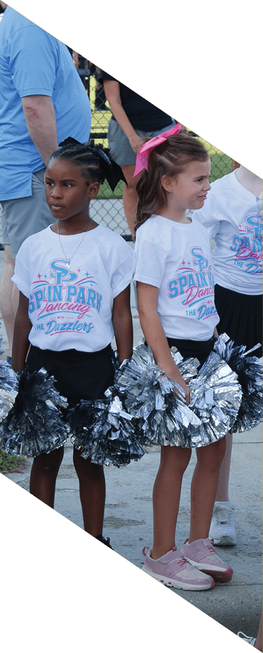 Children wearing matching dance team t-shirts and holding silver pom-poms stand on a sidewalk near a sports field. Description generated by AI