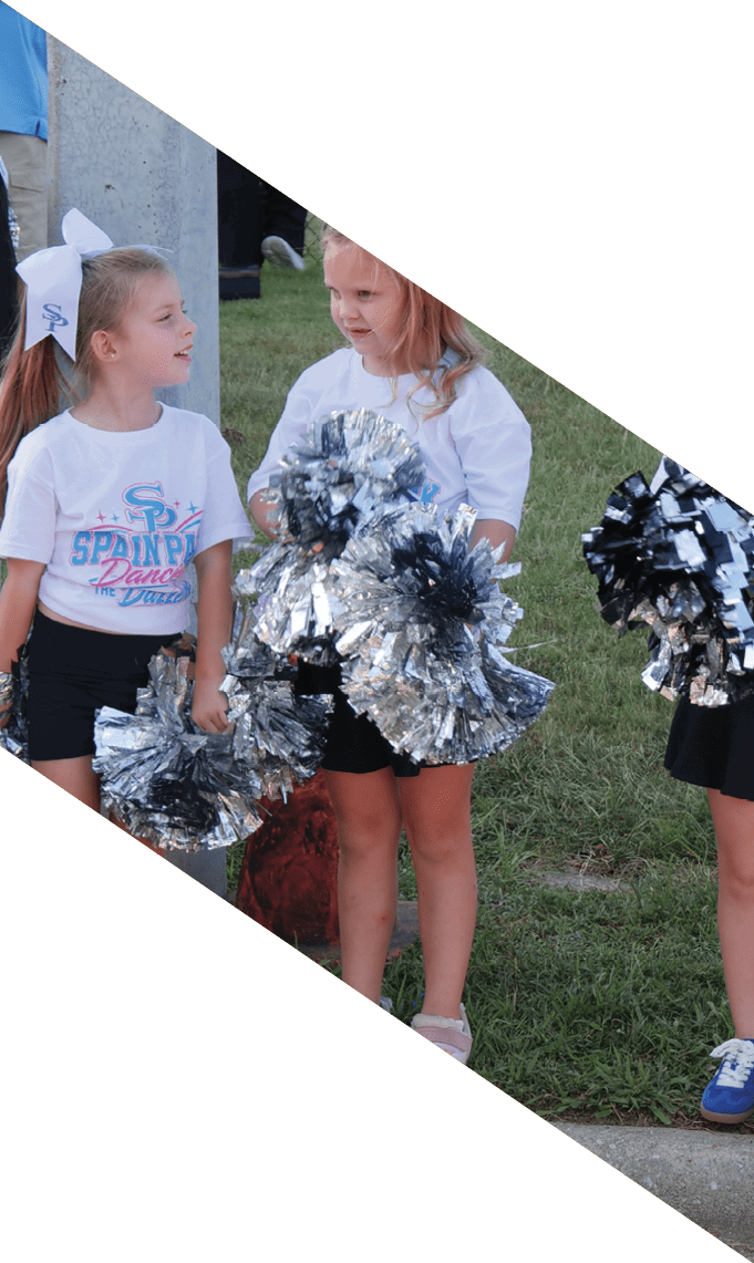 Children wearing matching cheerleading outfits with bows in their hair hold pom-poms while standing on grass. Description generated by AI