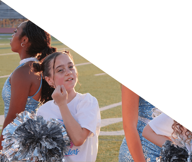 People in cheerleading outfits are standing on a sports field holding pom-poms, with one person in a white shirt making a gesture near their face. Description generated by AI