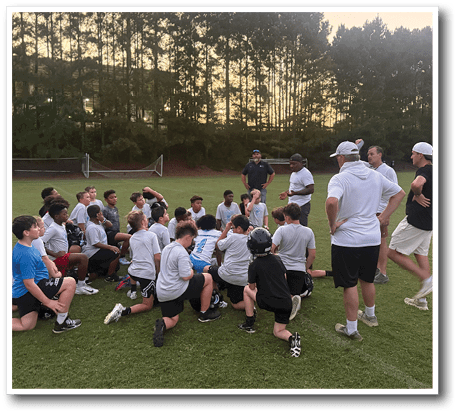 A group of people wearing sports attire are gathered on a grassy field with some kneeling and others standing while listening to a few adults who appear to be coaches during sunset. Description generated by AI