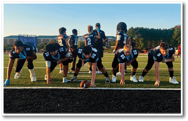 A group of people in football uniforms are lined up on a field in a ready stance with a football placed on the ground in front of them during sunset. Description generated by AI
