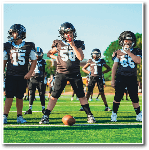 Several people in football uniforms stand on a green field with a football placed on the ground in front of them. Description generated by AI