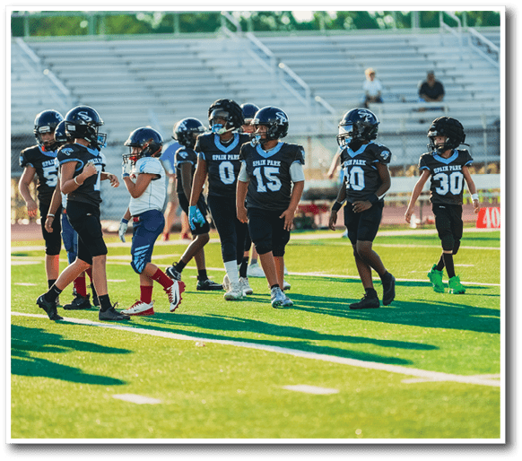 Children wearing football uniforms and helmets are standing and walking on a sunlit football field. Description generated by AI