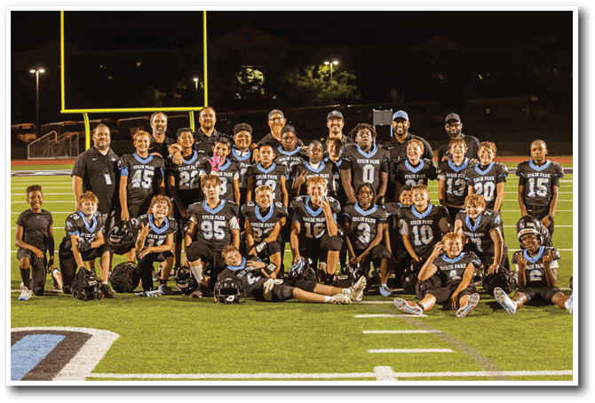 A group of children in football uniforms and adults in casual clothing pose together on a football field at night with a goalpost and lights in the background. Description generated by AI