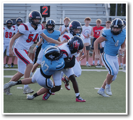 Several people wearing football uniforms are actively engaged in a tackle on a grassy field with others watching in the background. Description generated by AI