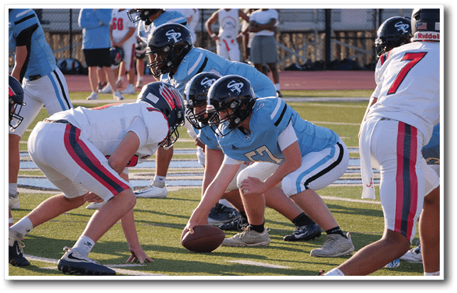 People in football uniforms are lined up on a field preparing for a play during a game. Description generated by AI