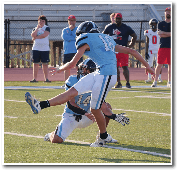 Two people in football uniforms are engaged in a play on a field while several people in casual and sports attire watch from the sidelines. Description generated by AI