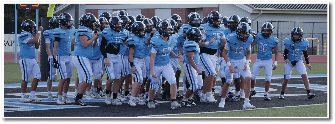 A group of people wearing light blue sports uniforms and helmets are gathered on a field near a goalpost with a digital play clock displaying the number 25. Description generated by AI