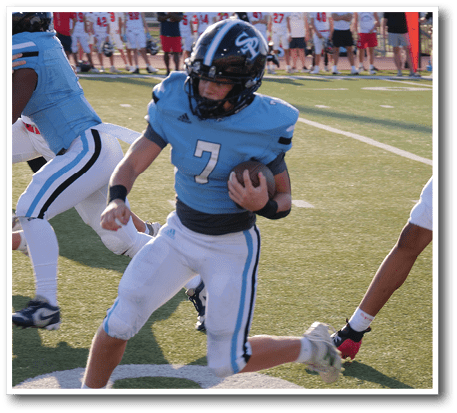 An adult wearing a blue football uniform with the number seven is running on a field while holding a football, with other players and spectators visible in the background. Description generated by AI