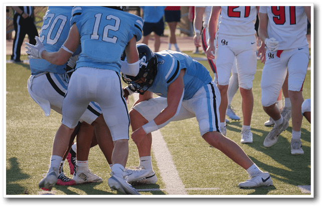 People in blue and white football uniforms are engaged in a play on a sunlit field with a white line marking the turf. Description generated by AI