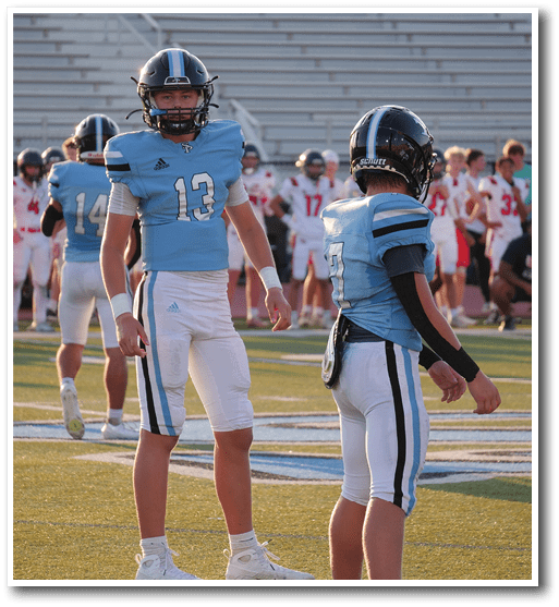 Two people in blue and white football uniforms stand on a field while others in red and white uniforms are visible in the background. Description generated by AI