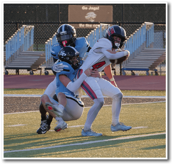 A person in white sports gear is running with a football while two people in blue uniforms attempt to tackle them on a football field. Description generated by AI