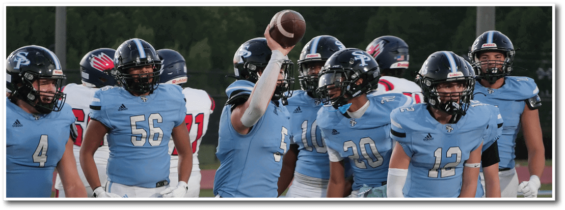 A group of people wearing light blue football uniforms and helmets are gathered on a field with one person holding a football above their head. Description generated by AI
