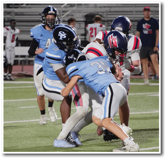 Players in blue uniforms are tackling a player in a white uniform with red and blue stripes during a football game on a field with spectators in the background. Description generated by AI