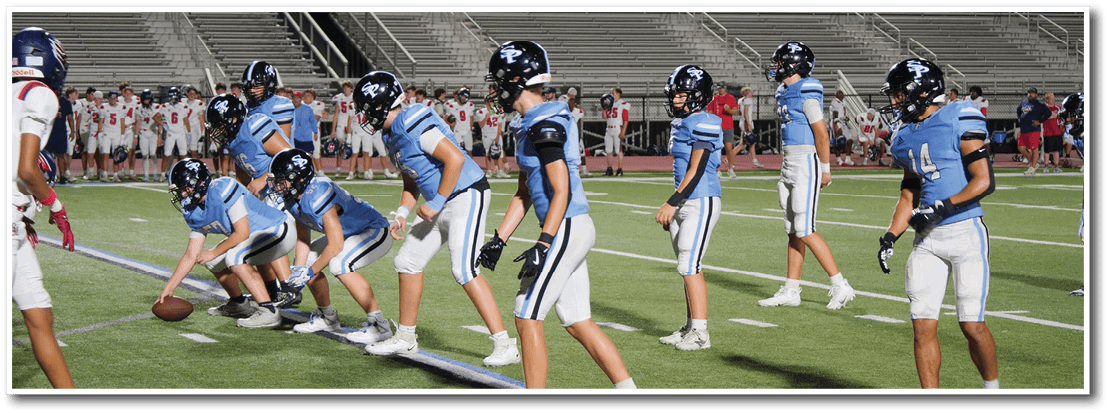 People in blue sports uniforms and helmets are lined up on a field with a sunset sky and empty bleachers in the background. Description generated by AI