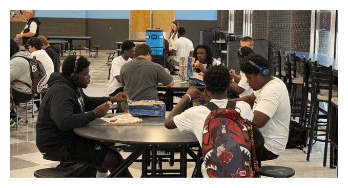 A group of people sit around a table in a cafeteria setting, some engaged in conversation while others appear to be eating or listening. Description generated by AI