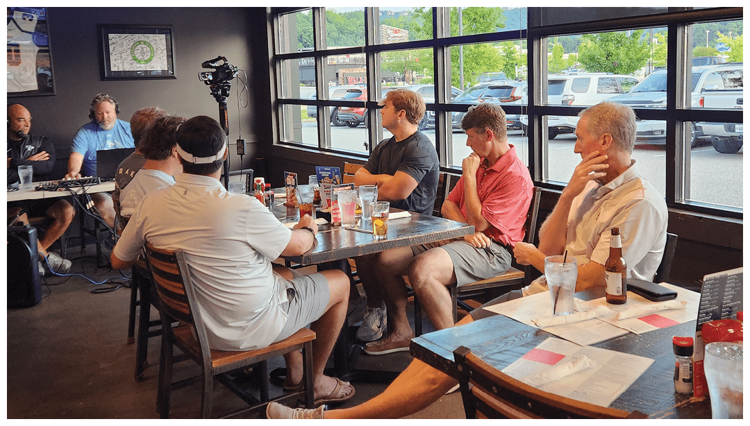 A group of adults sitting around a table in a casual restaurant with drinks and menus, engaged in conversation near large windows showing parked cars outside. Description generated by AI