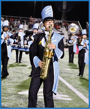 A person wearing a blue and black marching band uniform and hat is playing a saxophone on a field at night with other band members and a crowd in the background. Description generated by AI