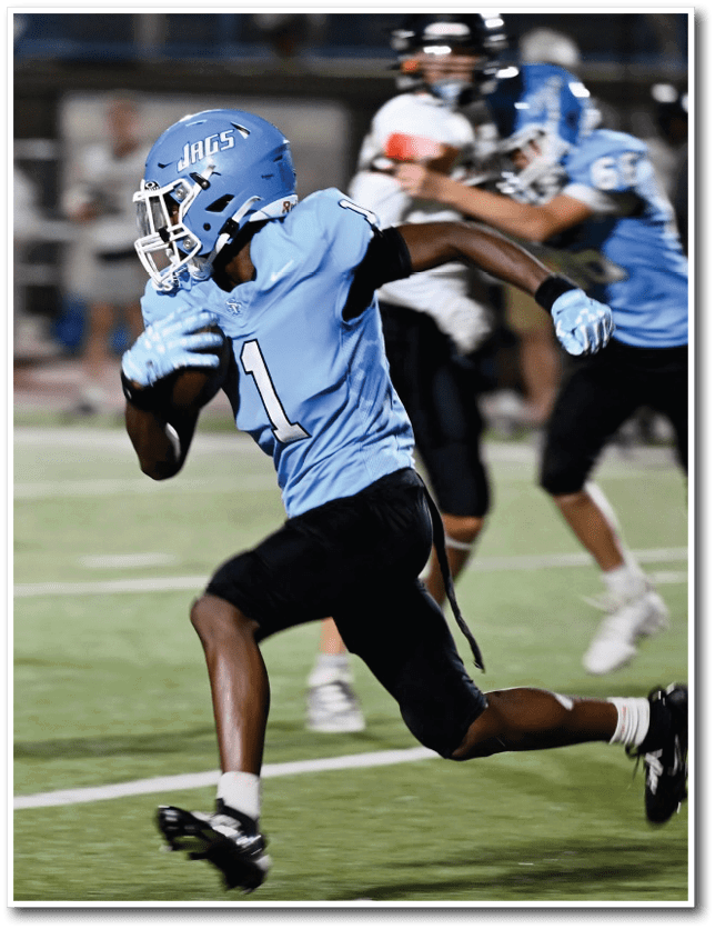 A person in a blue football uniform is running with the ball on a field while other players in blue and white uniforms are in the background. Description generated by AI