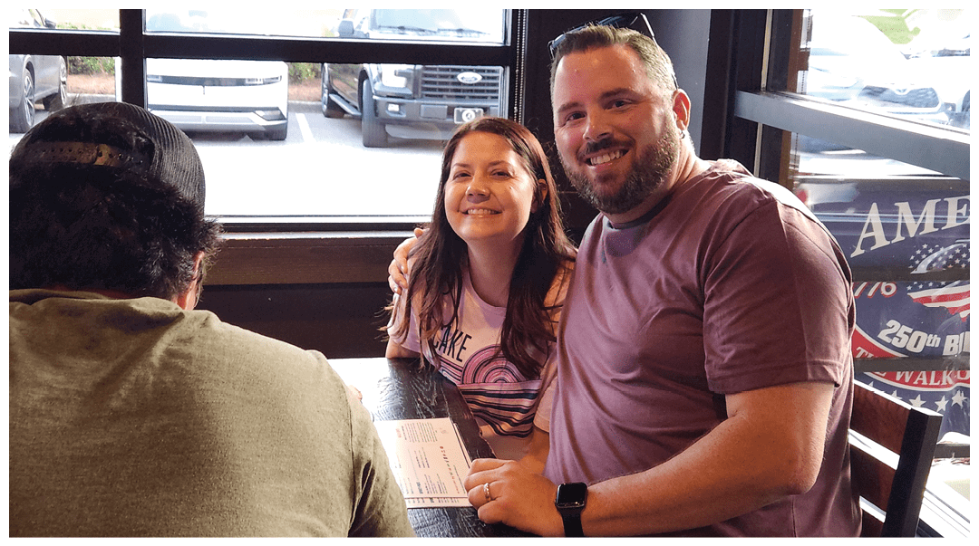 Three adults are sitting at a table in a casual dining setting with two of them smiling and looking toward the camera while the third has their back to the camera. Description generated by AI