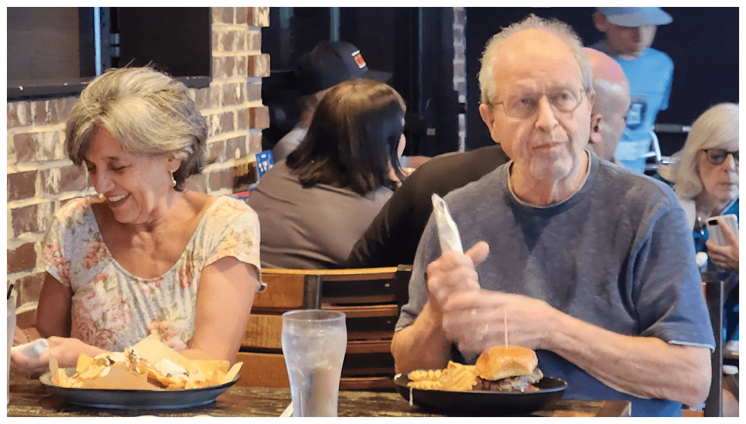 Two adults sit at a restaurant table smiling and eating sandwiches with drinks in front of them while other people are visible in the background. Description generated by AI