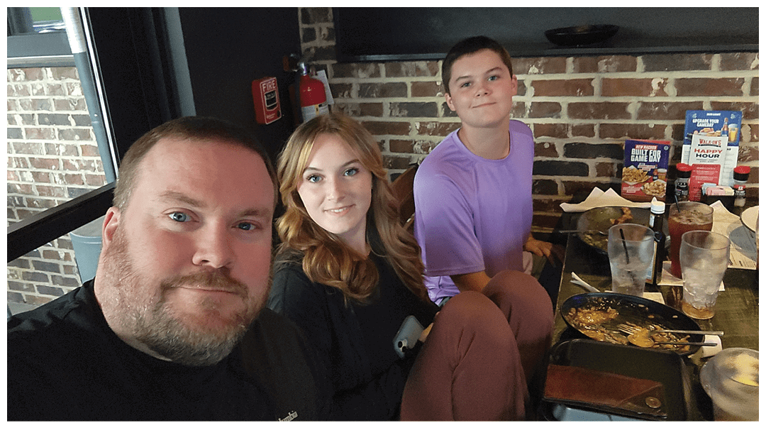 Three people sitting closely together at a restaurant table with finished plates and drinks, smiling at the camera. Description generated by AI