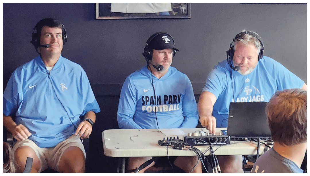 Three adults wearing headsets and blue Spain Park football shirts are seated at a table with audio equipment and a laptop, appearing to commentate or broadcast. Description generated by AI