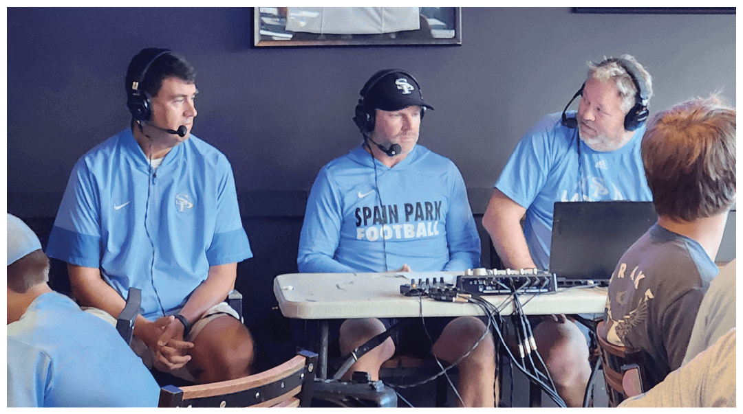 Three adults wearing headsets and light blue sports shirts are seated at a table with audio equipment, engaged in a discussion or broadcast. Description generated by AI