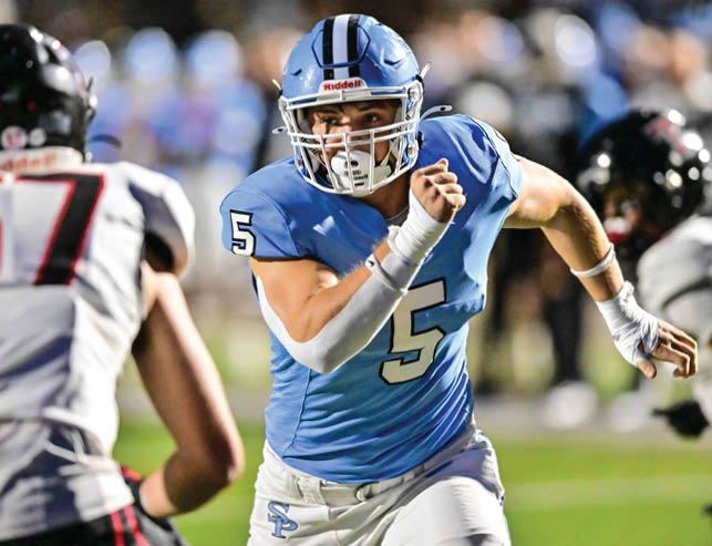 A person wearing a blue football uniform with the number 5 is running on a field while another player in a white and black uniform is nearby. Description generated by AI