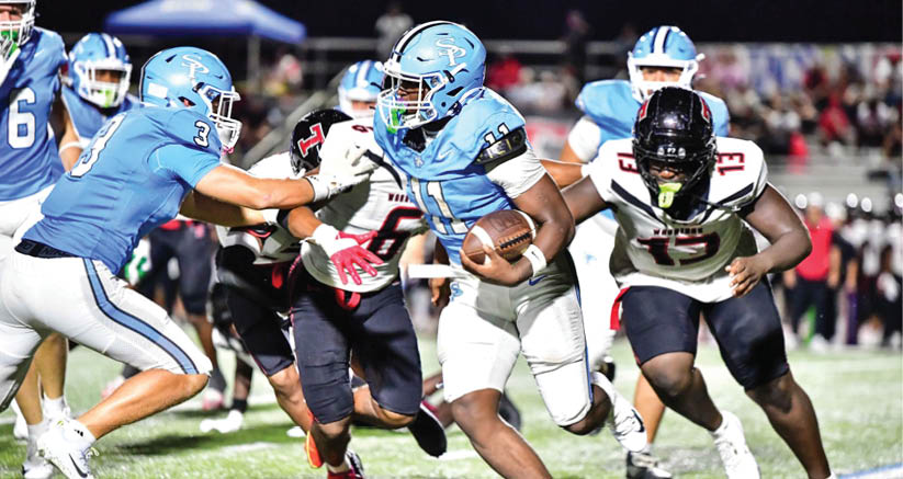 A group of people in football uniforms are actively playing on a field with one person in light blue running with the ball while others in light blue and black and white uniforms attempt to block or tackle. Description generated by AI