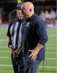 Two people stand on a sports field, one wearing a black and white striped shirt and the other in dark athletic clothing, with the person in dark clothing gesturing with their hand. Description generated by AI