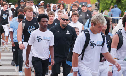 A group of people wearing sports attire and backpacks walk together outdoors near steps, with some wearing white shirts labeled “Jaguars." Description generated by AI