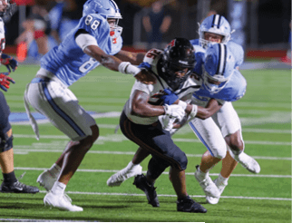 Three football players in light blue uniforms are tackling a player in a black and white uniform who is holding the ball on a green field. Description generated by AI