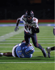 A person in a light blue football uniform is on the ground while a person in a white and dark uniform runs past them on a football field. Description generated by AI