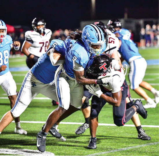 People wearing football uniforms are engaged in a tackle on a brightly lit field during a night game. Description generated by AI