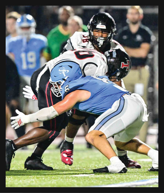 Two people in football uniforms are engaged in a tackle on a grassy field during a game, with one player in blue and white gear reaching out to stop the player in black and white gear holding the ball. Description generated by AI