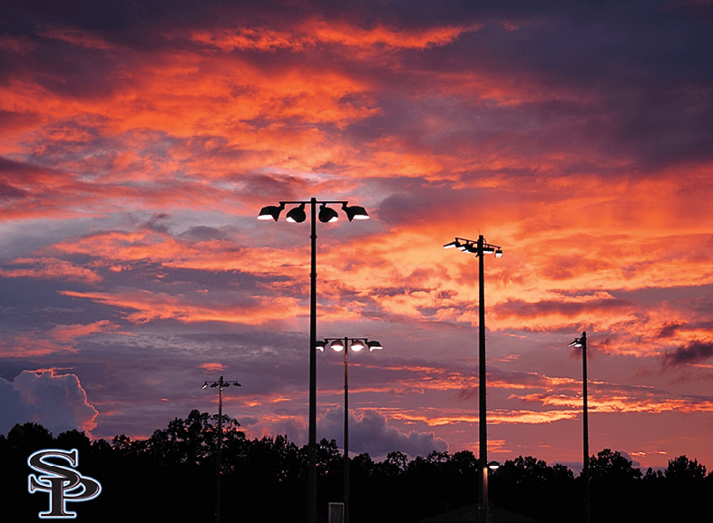 A vibrant orange and purple sunset sky with silhouetted tall streetlights and trees in the foreground. Description generated by AI