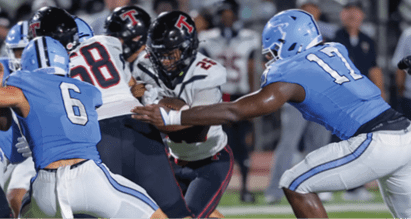 Two people in blue football uniforms are attempting to block a person in a white and black football uniform who is running with the ball on a grassy field. Description generated by AI