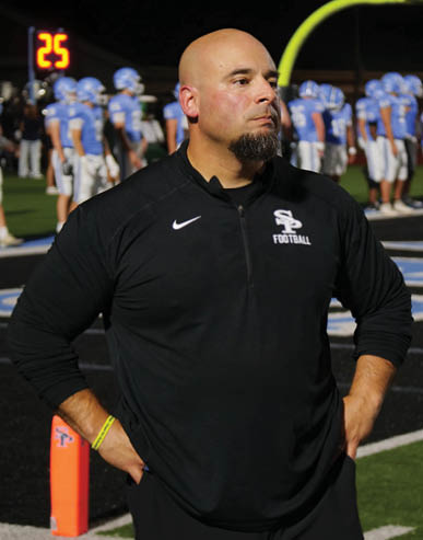A person wearing a black sports jacket with a football logo stands on a football field with players in blue uniforms in the background. Description generated by AI