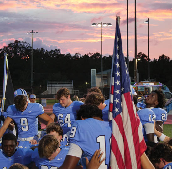 A group of people wearing blue football uniforms are gathered on a field near an American flag with a sunset sky and stadium lights in the background. Description generated by AI
