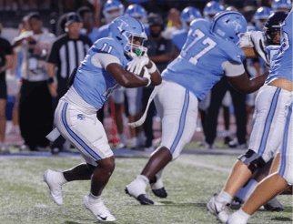 Several people in light blue football uniforms are actively playing on a grassy field during a game. Description generated by AI